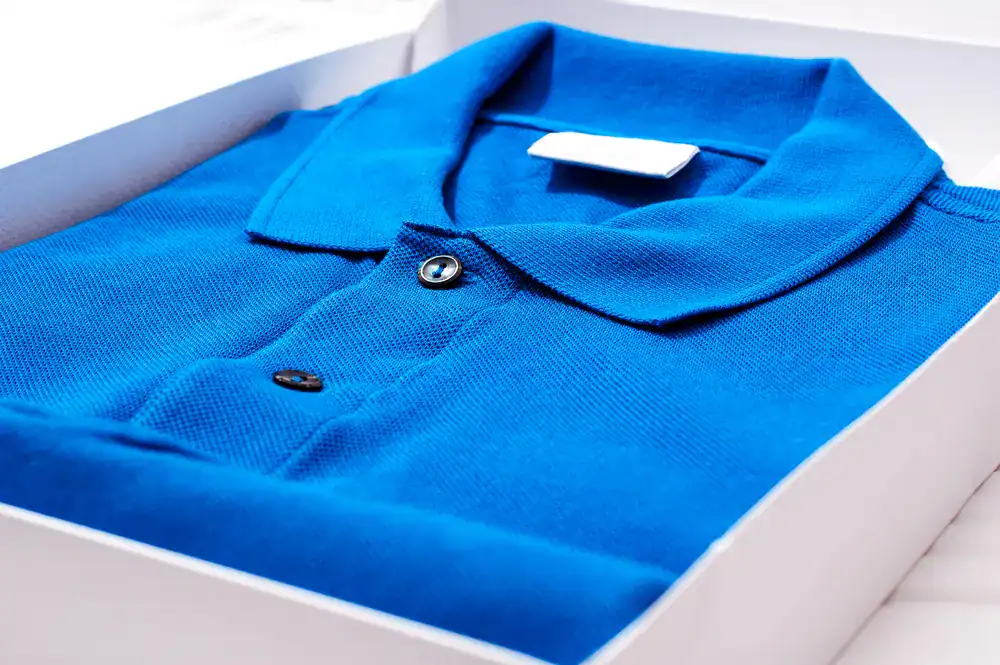 A neatly folded blue polo shirt with black buttons is placed inside a white box, with a blank white tag on the collar.