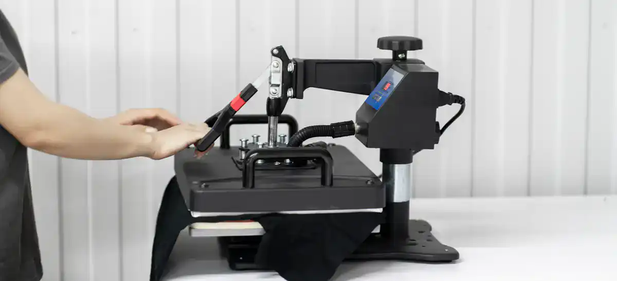 A person operates a heat press machine, pressing a black fabric item onto the heated plate, with their hand adjusting the handle. The background features a white paneled wall.